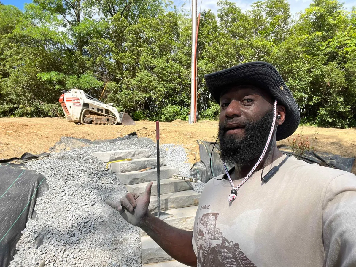 Otis Thomas on a job site beside newly set natural stone steps, with a compact track loader behind him